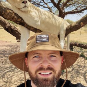 A man with a hat takes a selfie under a white lion resting on a tree in Kilimanjaro, Tanzania.